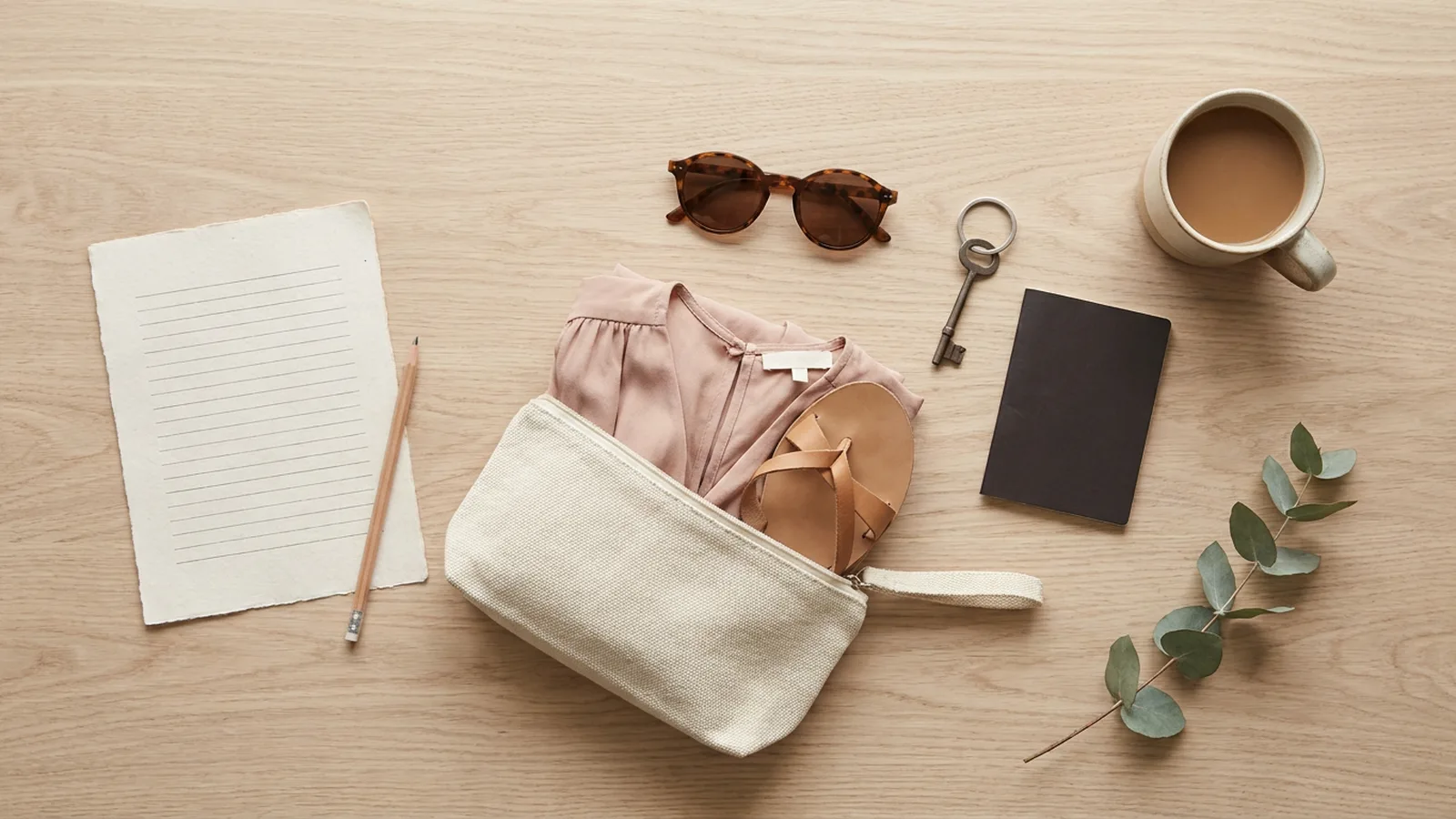 Illustration: a blank lined sheet and pencil, a canvas pouch with a pink top and sandals, sunglasses, a key, a notebook and coffee on a wooden table.